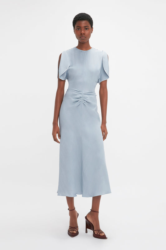 A woman wearing Victoria Beckham's Exclusive Gathered Waist Midi Dress In Pebble, a light blue fit-and-flare silhouette midi dress with cap sleeves and a front tie, standing against a white background.