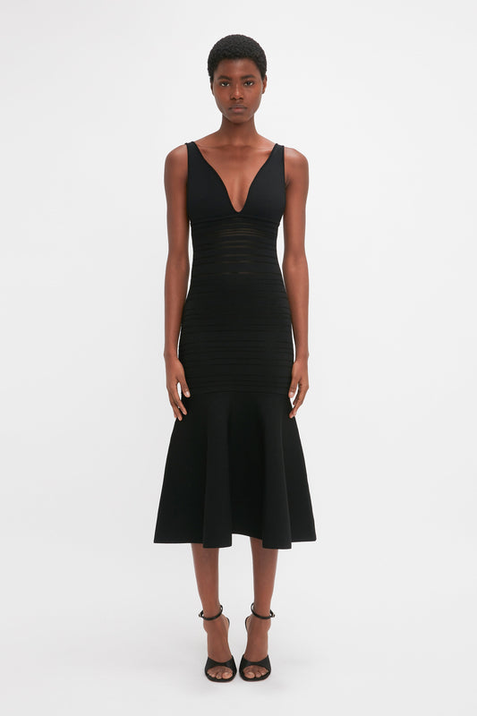 A black woman stands facing the camera, wearing a fitted black Victoria Beckham Frame Detail Sleeveless Dress with a v-neckline and midi-length hemline, paired with black heeled sandals.