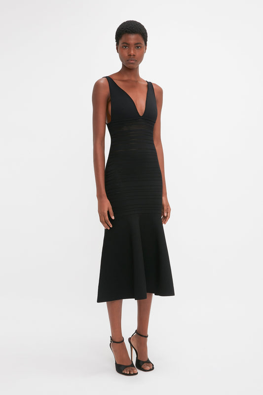 A black woman in a Victoria Beckham black sleeveless fit-and-flare dress with a plunging neckline and midi-length hemline, posing against a white background.