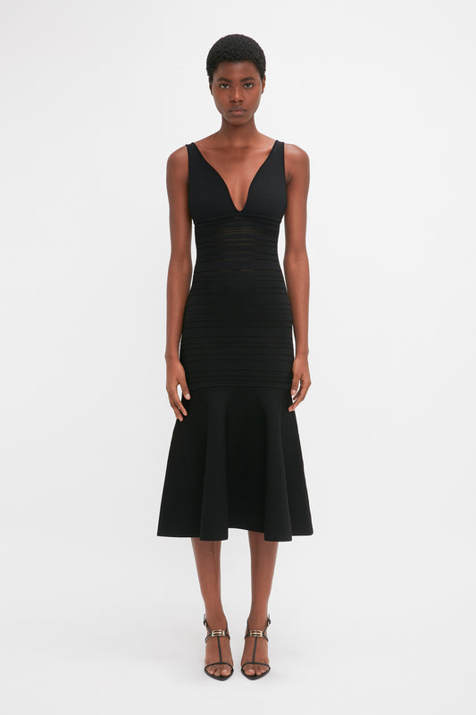 Person standing against a plain white background, wearing a form-fitting sleeveless black dress with a V-neck and hemline flares, paired with Victoria Beckham Frame Detail Sandal In Black Leather featuring sculptural heel.