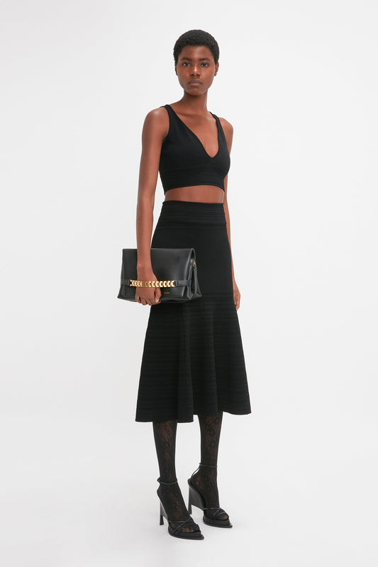 A person stands wearing a black crop top and a waist-defining Victoria Beckham Fit And Flare Midi Skirt In Black, holding a black clutch bag. They complement their outfit with black tights and high-heeled shoes, while the contrasting stitching adds an extra touch of elegance.