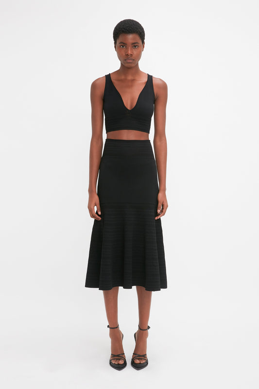 A person stands against a plain white background, wearing a sleeveless black top and the Victoria Beckham Fit And Flare Midi Skirt In Black with flared hem. The black high-heeled shoes complement the outfit perfectly, showcasing subtle contrasting stitching for added elegance.