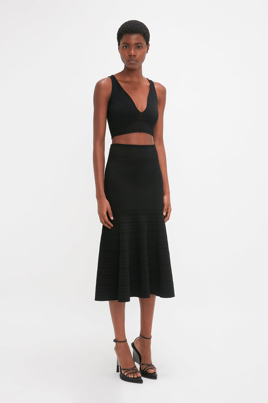 A person wearing a black sleeveless crop top and a waist-defining, mid-weight stretch Fit And Flare Midi Skirt In Black by Victoria Beckham stands against a white background. They are also wearing high-heeled sandals.