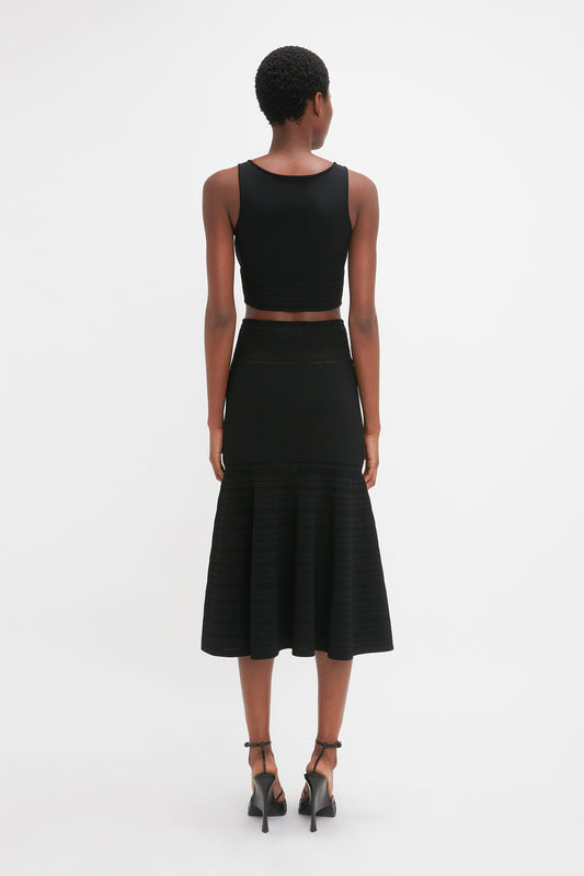 A person stands facing away from the camera, wearing a black sleeveless crop top and a Victoria Beckham Fit And Flare Midi Skirt In Black, made of mid-weight stretch knit material. They complete their ensemble with black high-heeled shoes against a plain white background.