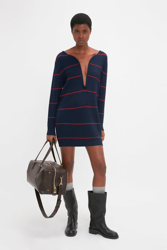 Person standing against a white background, wearing the Victoria Beckham Frame Detail Jumper Dress In Navy-Red, black boots, and holding a brown duffle bag.