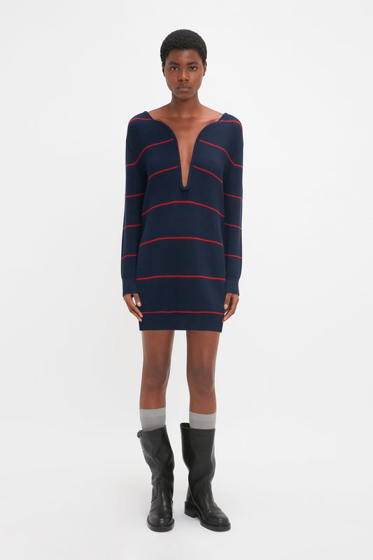 A person stands facing forward, wearing a Victoria Beckham Frame Detail Jumper Dress In Navy-Red with a directional curved neckline, black knee-high boots, and gray socks. The background is plain white.