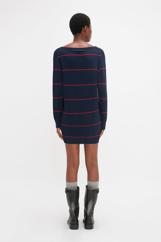 A person is standing and facing away, wearing a navy blue, long-sleeved, striped Frame Detail Jumper Dress In Navy-Red from Victoria Beckham's SS24 runway and knee-high black boots with gray socks showing above the boots.