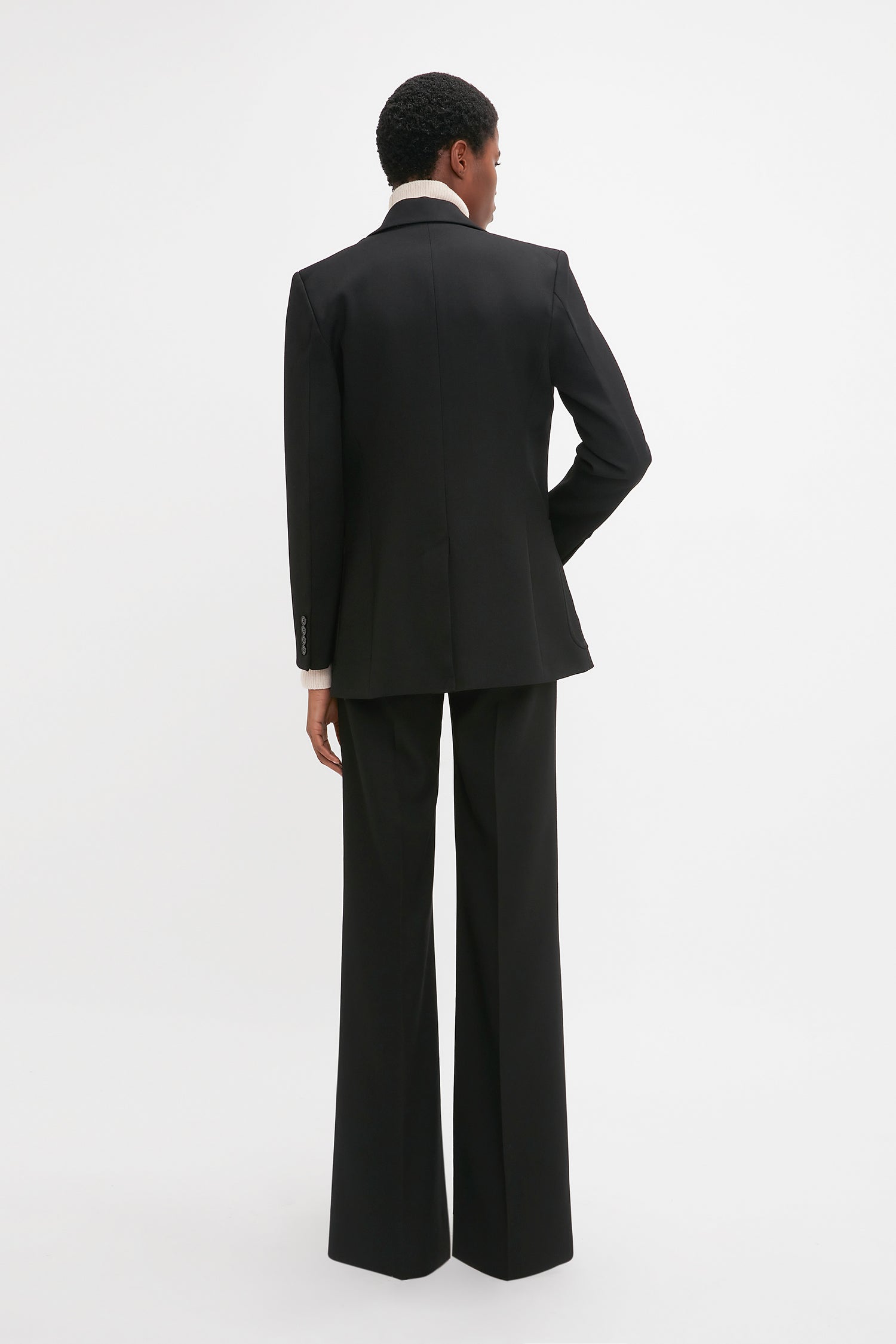 A person stands with their back to the camera, wearing a modern Patch Pocket Jacket In Black by Victoria Beckham with wide-leg trousers and a white shirt. The background is plain white.