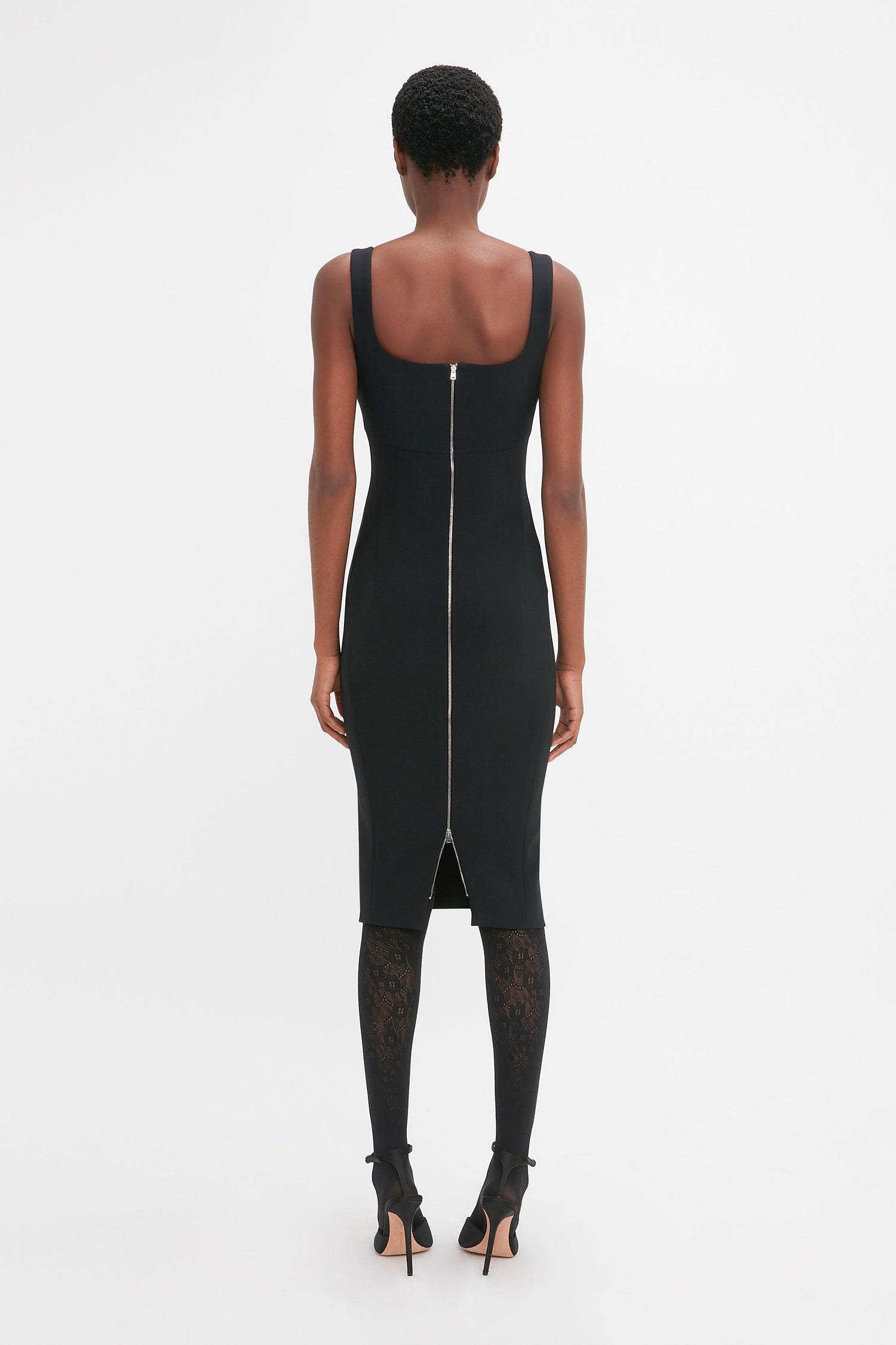 A person stands facing away, wearing the Victoria Beckham Sleeveless Fitted T-Shirt Dress In Black with an exposed double-ended zip down the back. The knee-length black dress is complemented by patterned black stockings and high-heeled shoes.