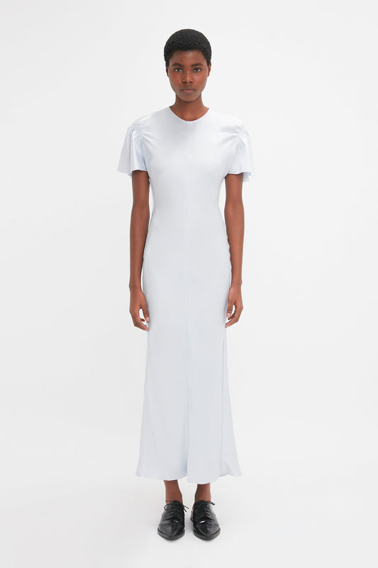 A person stands against a plain white background, wearing the Victoria Beckham Gathered Sleeve Midi Dress In Ice with short sleeves and black lace-up shoes. The light gray fabric drapes beautifully, featuring a subtle godet insert that adds volume and movement to the garment.