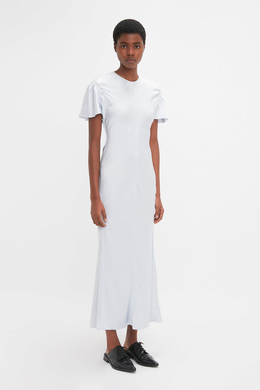 A person with short hair is standing against a plain white background, wearing an elegant Gathered Sleeve Midi Dress In Ice by Victoria Beckham and black shoes.