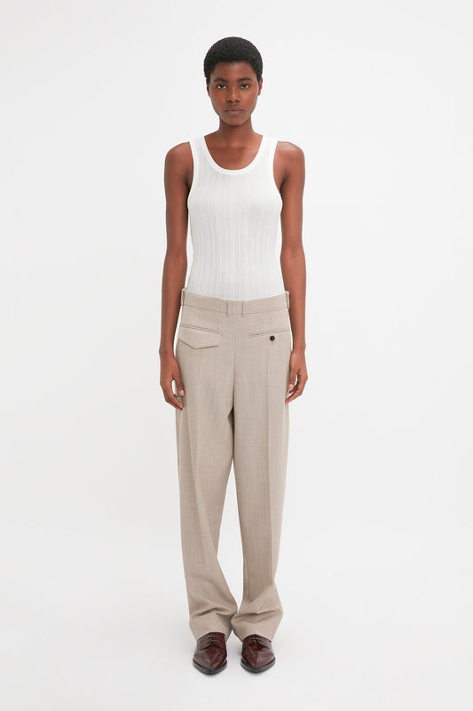 A modern woman is wearing a white sleeveless top and Victoria Beckham's Reverse Front Trouser In Sesame, paired with brown shoes, standing against a plain white background.