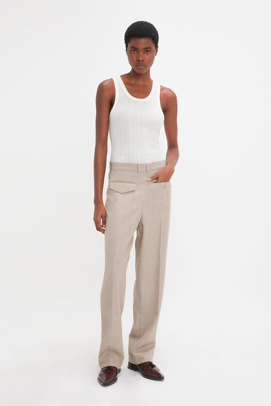 A person with short hair is wearing a white, Fine Knit Vertical Stripe Tank In White by Victoria Beckham and beige trousers, standing against a plain white background, exuding a subtle yet elegant textured appearance.
