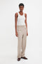 Person wearing a Fine Knit Vertical Stripe Tank In White by Victoria Beckham, beige high-waisted trousers, and brown shoes, standing against a plain white background. The outfit's textured appearance evokes effortless elegance reminiscent of Victoria Beckham's style.