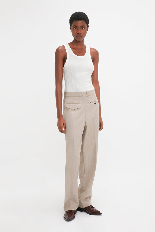 A person stands against a plain background wearing a white tank top, Victoria Beckham Reverse Front Trouser In Sesame, and brown shoes.