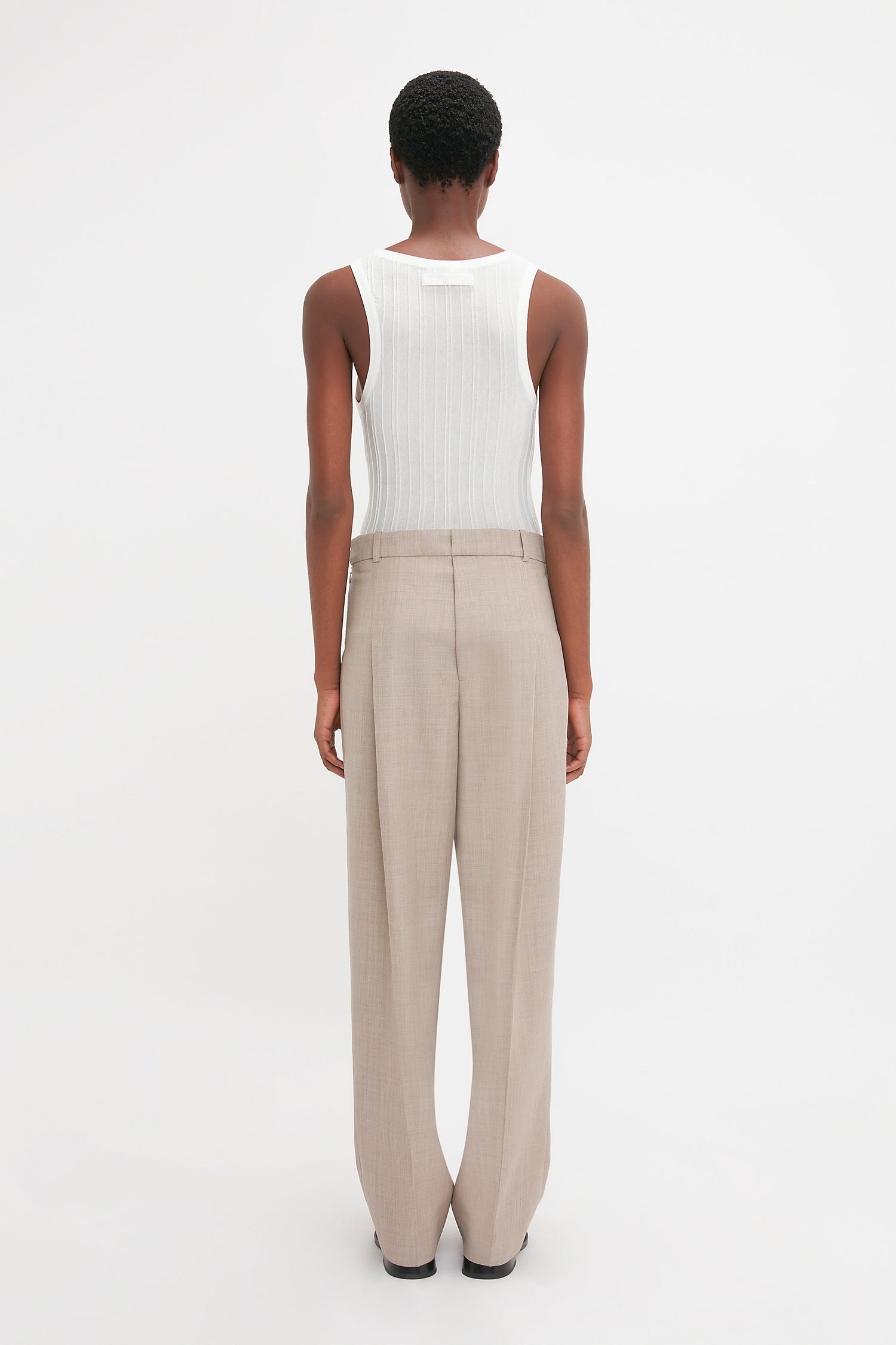 Person standing, facing away, wearing a Fine Knit Vertical Stripe Tank In White and beige trousers against a plain white background. The ensemble, reminiscent of Victoria Beckham's minimalist style, boasts a subtly textured appearance that adds depth to the simplicity.