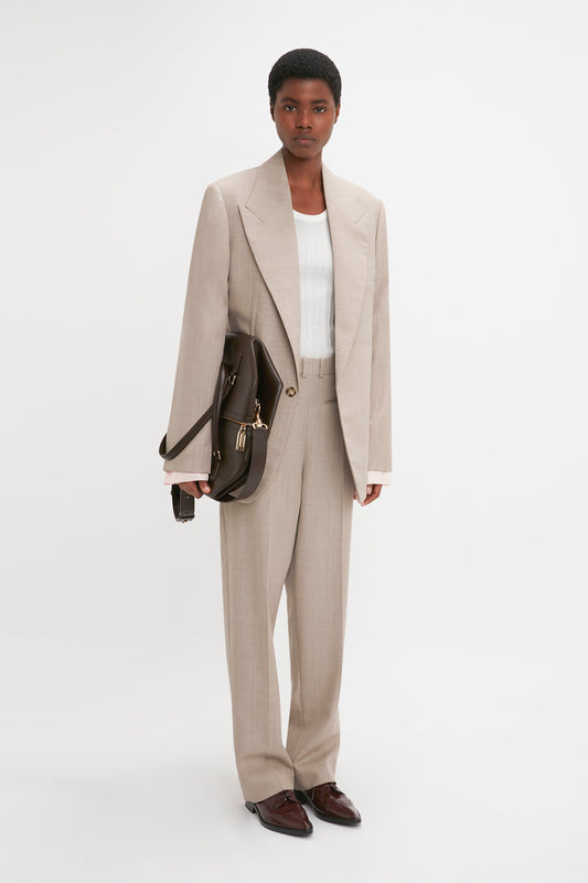 A person poses in a light beige Darted Sleeve Tailored Jacket In Sesame by Victoria Beckham made from pure wool, paired with a white top, brown shoes, and a brown satchel bag against a plain white background.