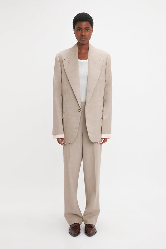 A person stands against a plain background, wearing an oversized beige suit made of pure wool, accompanied by a white shirt and brown shoes. The Darted Sleeve Tailored Jacket In Sesame by Victoria Beckham adds contemporary detailing to the classic ensemble.