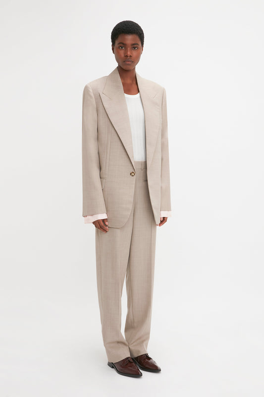 A person wearing a beige pure wool suit with a Darted Sleeve Tailored Jacket In Sesame by Victoria Beckham, complemented by a white shirt and brown shoes, stands against a white background.