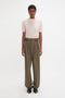 A person wearing the Victoria Beckham Short Sleeve Top In Nougat and high-waisted, wide-legged olive-green trousers stands against a plain white background.