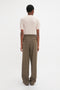 Person with short curly hair wearing a Short Sleeve Top In Nougat by Victoria Beckham and high-waisted, loose-fitting brown pants, featuring a delicate pointelle lace stitch pattern, standing with their back facing the camera against a plain white background.