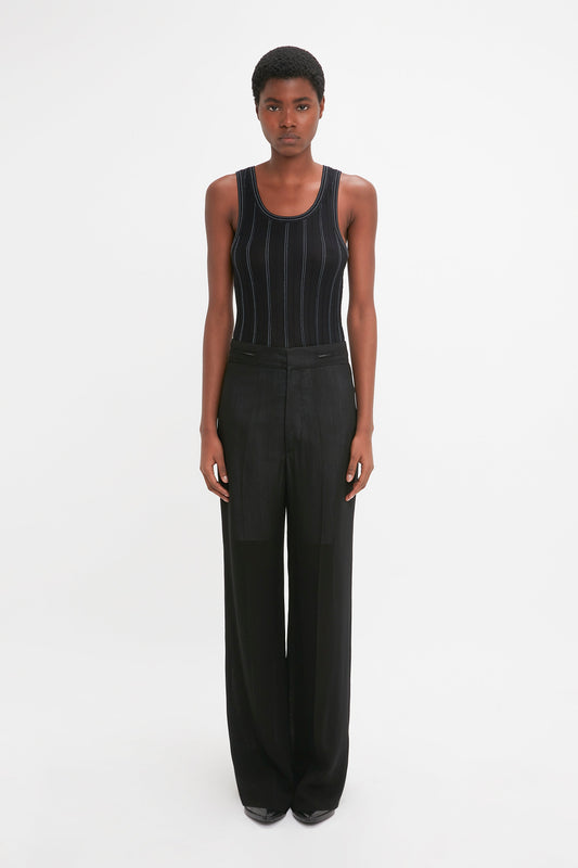 Person standing against a white background, wearing a black Fine Knit Vertical Stripe Tank In Black-Blue by Victoria Beckham and black wide-leg trousers.