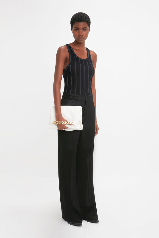 A person stands against a plain white backdrop wearing a black Fine Knit Vertical Stripe Tank In Black-Blue by Victoria Beckham and matching pants, holding a white clutch bag with a gold chain.