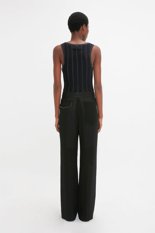 Person standing facing away from the camera, wearing a Fine Knit Vertical Stripe Tank In Black-Blue and black pants against a plain white background, resembling the sleek elegance of Victoria Beckham’s style.