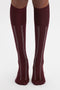 A person wears knee-high burgundy and white socks with thin vertical stripes, standing on a white surface. Crafted from 100% cotton, these Superfine Rib Socks In Burgundy by Victoria Beckham offer both comfort and style.