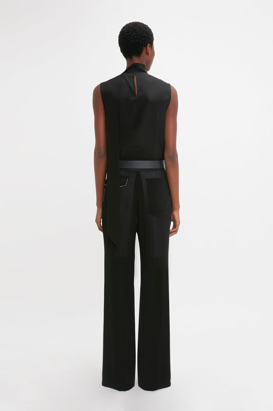 A person with short hair stands facing away, wearing a Victoria Beckham Sleeveless Tie Neck Top In Black and high-waisted black pants with a belt, set against a plain white background.