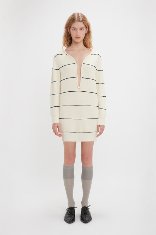 Young person with long hair wearing a Victoria Beckham Frame Detail Jumper Dress In Natural-Navy featuring a V-shape neckline, knee-high socks, and black shoes, standing against a plain white background.