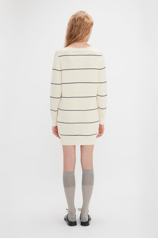 A person with long, wavy hair is standing with their back to the camera, wearing a Victoria Beckham Frame Detail Jumper Dress In Natural-Navy, gray knee-high socks, and black shoes, against a plain white background.