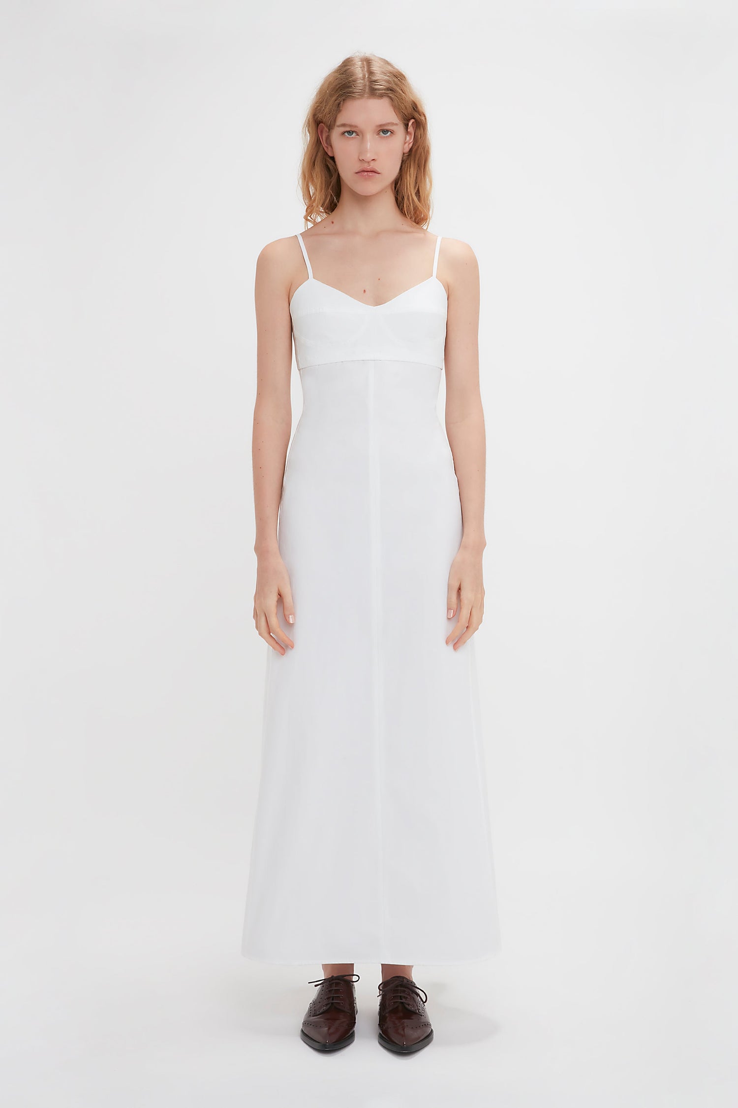 A woman in a long white Victoria Beckham Cami Fit And Flare Midi In White stands against a plain white background, facing forward with a neutral expression. Her outfit exudes a subtle bohemian vibe, complemented by dark brown shoes.