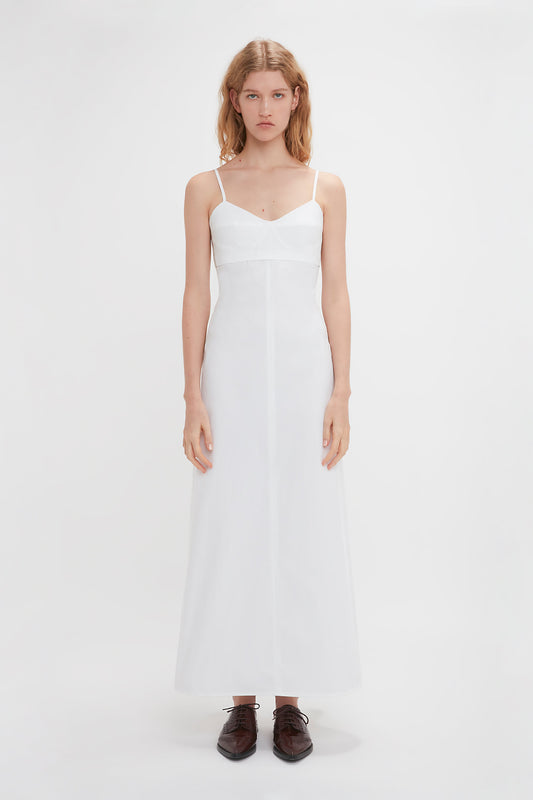 A woman in a long white Victoria Beckham Cami Fit And Flare Midi In White stands against a plain white background, facing forward with a neutral expression. Her outfit exudes a subtle bohemian vibe, complemented by dark brown shoes.