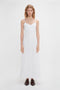 A woman in a long white Victoria Beckham Cami Fit And Flare Midi In White stands against a plain white background, facing forward with a neutral expression. Her outfit exudes a subtle bohemian vibe, complemented by dark brown shoes.