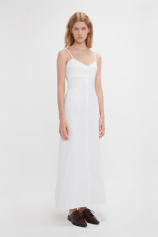 A woman stands against a white background, wearing the Victoria Beckham Cami Fit And Flare Midi In White with thin straps and black dress shoes.