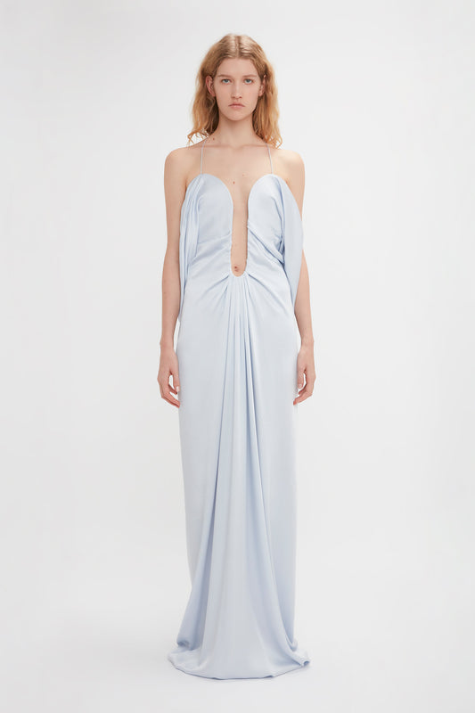 A person standing and wearing a light blue, floor-length crepe back satin dress with a deep V-neckline and thin straps, set against a plain white background. Perfect eveningwear, this Frame Detail Cut-Out Cami Dress In Ice by Victoria Beckham exudes elegance.