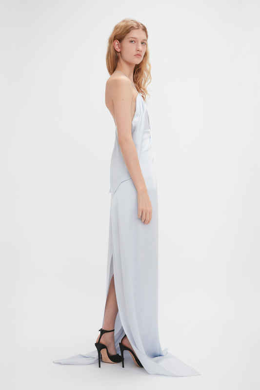A person with long hair stands in a Victoria Beckham Frame Detail Cut-Out Cami Dress In Ice and black high heels, facing the camera with a neutral expression against a plain white background.