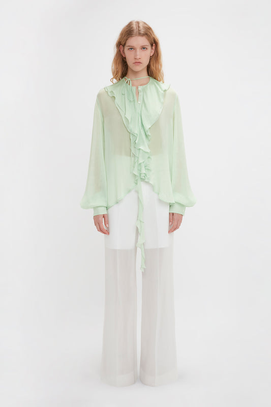 A person stands against a white background wearing the Romantic Blouse In Jade by Victoria Beckham and white, semi-sheer wide-leg pants.