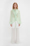 A person stands against a white background wearing the Romantic Blouse In Jade by Victoria Beckham and white, semi-sheer wide-leg pants.