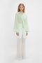 Young woman modeling the Victoria Beckham Romantic Blouse In Jade, a sheer light green hue with blouson sleeves and ruffles, paired with flowing white trousers against a plain white background.