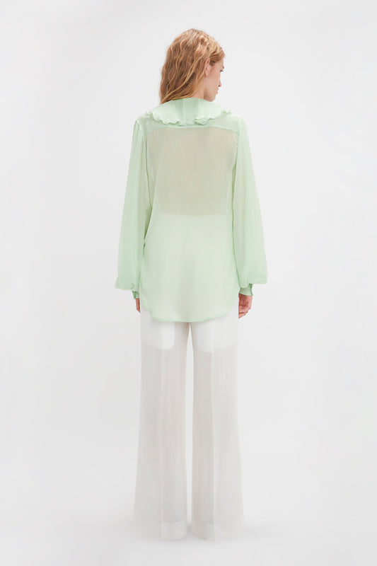 A person with light-colored hair is seen from the back, wearing a semi-sheer, light green Romantic Blouse In Jade by Victoria Beckham with blouson sleeves and white wide-leg pants against a plain white background.