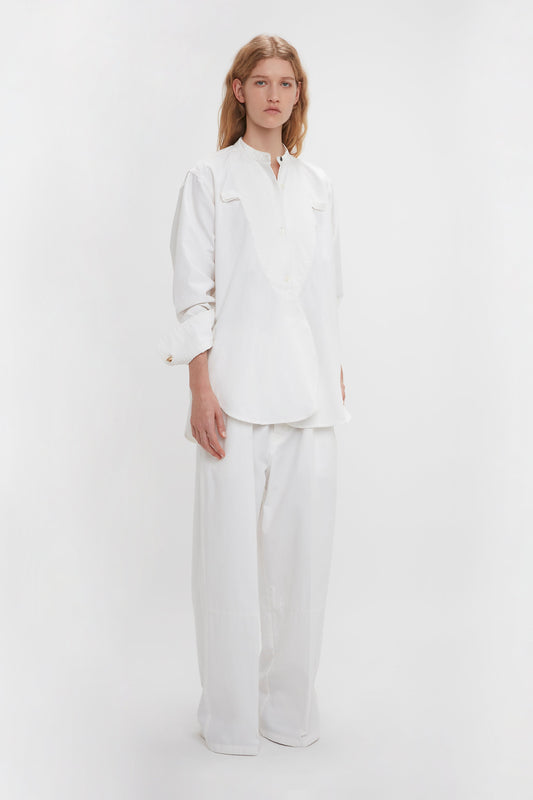 A person with long hair is wearing a relaxed fit, loose-fitting Bib-Front Tuxedo Shirt In Washed White by Victoria Beckham with a grandfather collar and wide-legged white pants, standing against a plain white background.