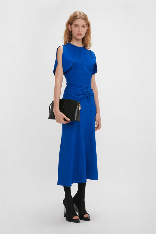A woman in a Victoria Beckham Gathered Waist Midi Dress In Palace Blue with cap sleeves and a black handbag stands against a white background.