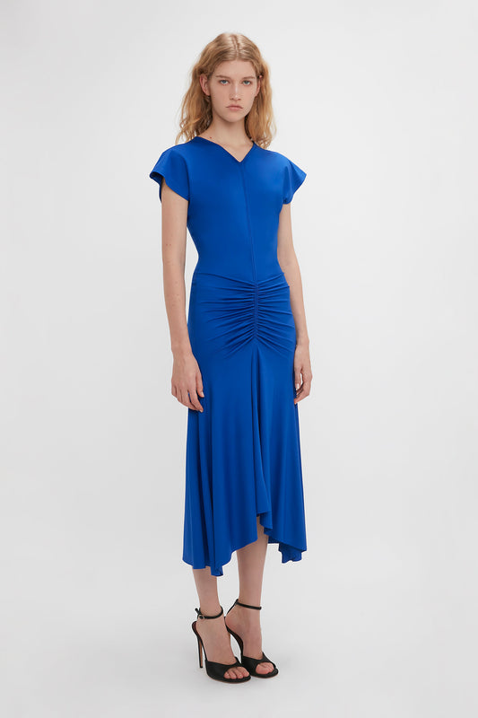 A woman in a Victoria Beckham bright royal blue Sleeveless Rouched Jersey Dress with cap sleeves stands against a white background.