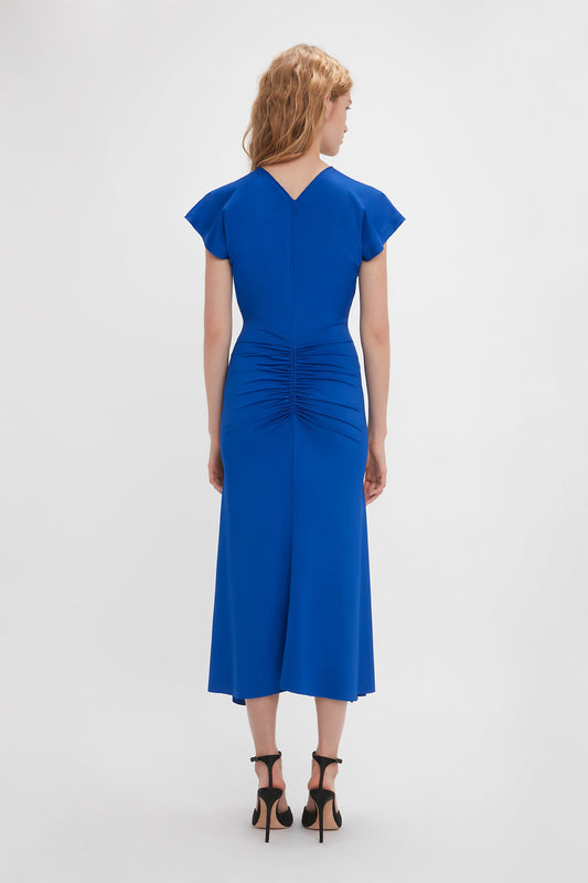 A woman in a Victoria Beckham royal blue midi dress with short sleeves and ruched detailing at the back, standing against a white background. She is viewed from the back.