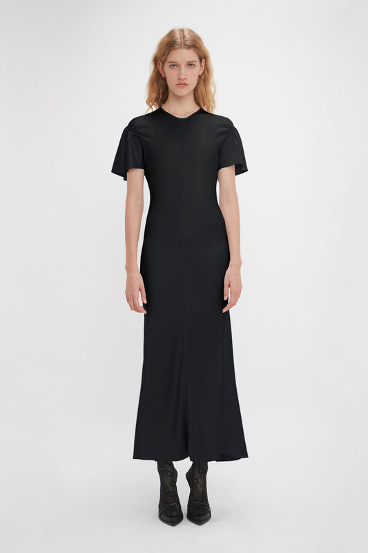 A person stands in a long, Gathered Sleeve Midi Dress In Black by Victoria Beckham made of crepe back satin and black shoes, against a plain white background.