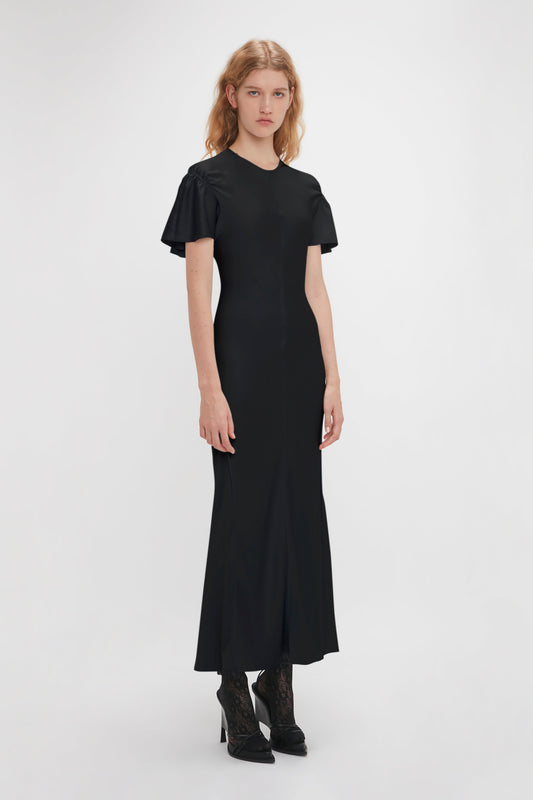 A woman with long blonde hair is wearing the Gathered Sleeve Midi Dress In Black by Victoria Beckham and black lace-up shoes, standing against a plain white background.