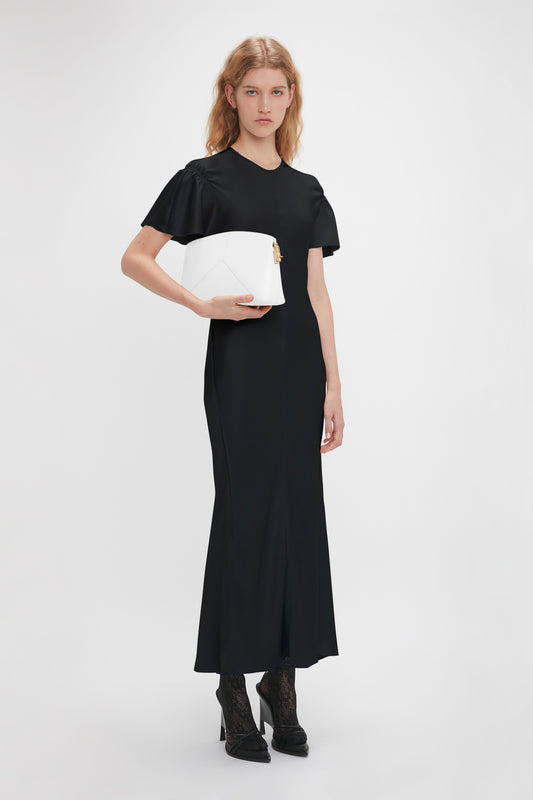 A woman with long blonde hair wearing a black floor-length dress and black shoes holds an Exclusive Victoria Clutch Bag In White Leather by Victoria Beckham against a plain white background.