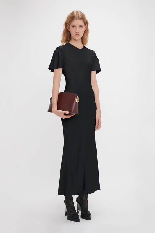 A woman with long blonde hair, reminiscent of Victoria Beckham's style, wears a black dress and black heeled shoes, clutching a Victoria Clutch Bag In Burgundy Leather by Victoria Beckham. She stands against a plain white background.
